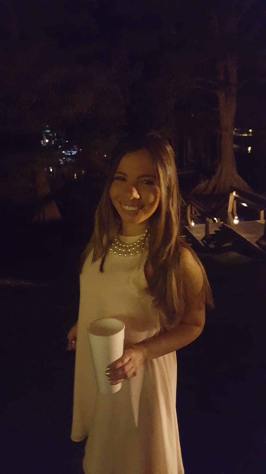 Silvana in a cream dress and pearl necklace at night, warm lights and water behind her.