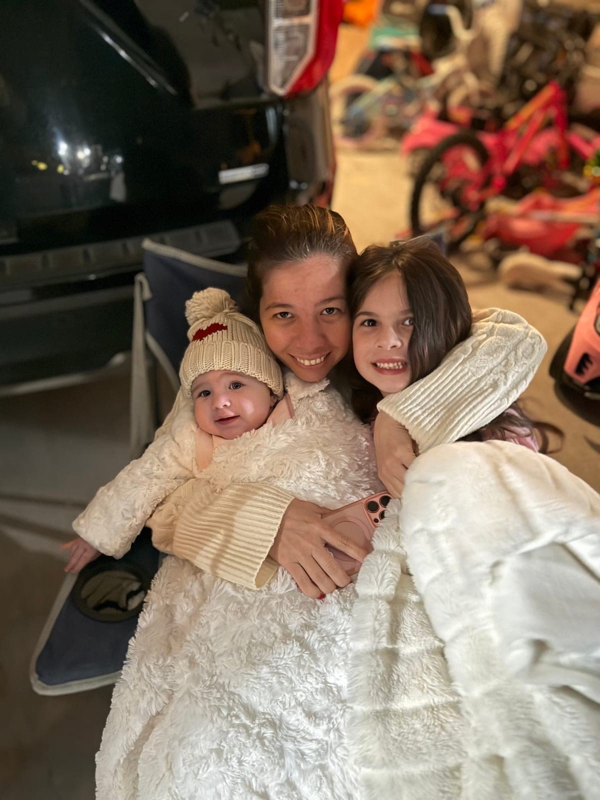 Silvana wrapped in a cream blanket in the garage, holding her baby in a knit bobble hat with her older daughter tucked under her arm.