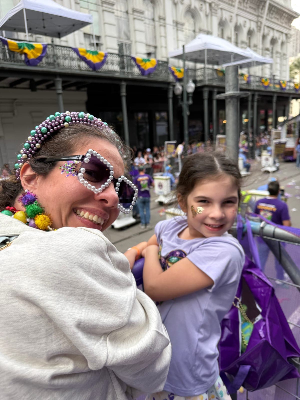 Silvana in pearl heart sunglasses and a beaded headband at the parade rail with her daughter, both grinning.