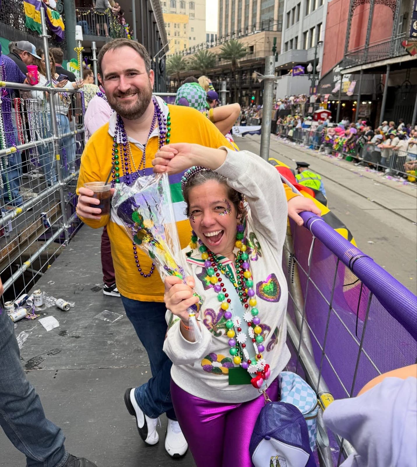 Silvana laughing at a Mardi Gras parade barricade in a beaded sweatshirt and magenta pants, Shaun behind her in a gold jersey.